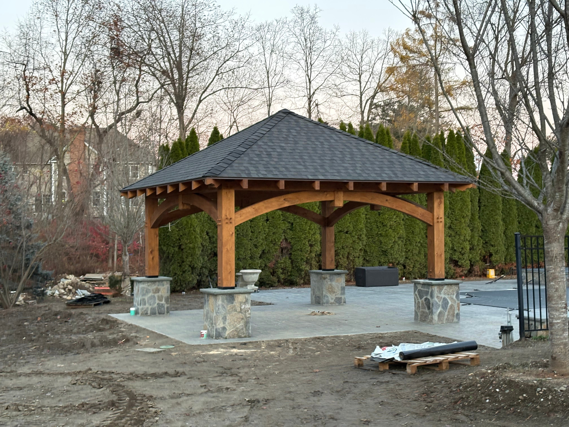 Finished timber frame pavilion with shingled hip roof and stone column bases on a paver patio